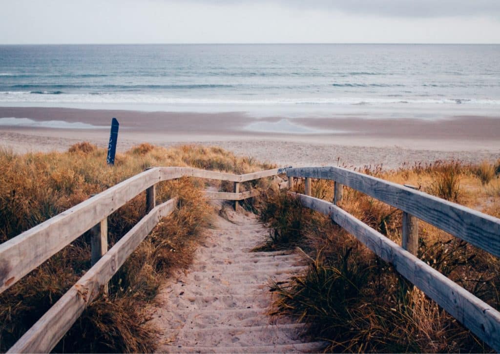 Wooden boardwalk pathway leading through sand dunes to a quiet beach with ocean waves under a cloudy sky in Cedar Point, NC